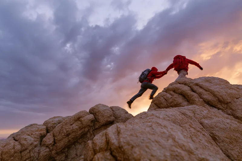 Two hikers helping each other climb rocky cliff at sunset