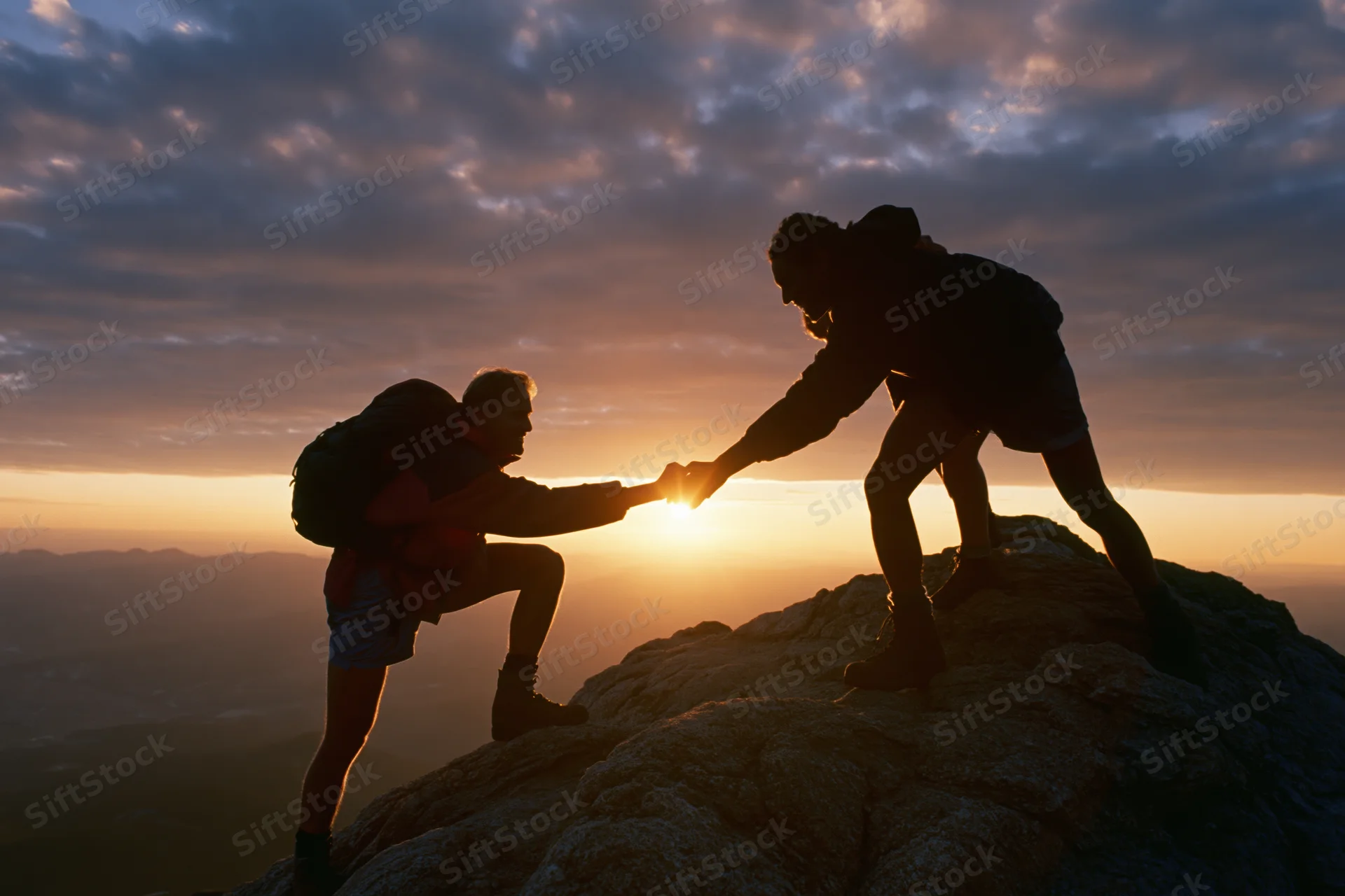 Two hikers silhouetted against sunset on mountain peak, one reaching down to help the other climb up