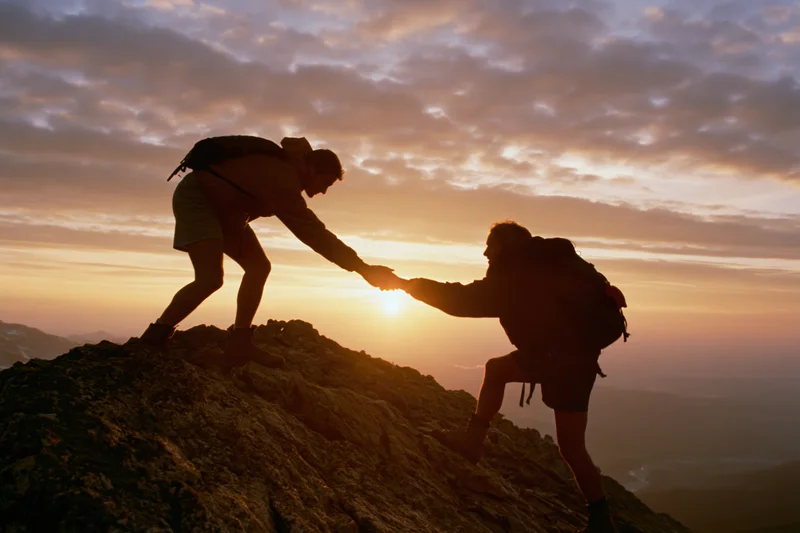 Two hikers silhouetted against sunset reaching to help each other climb rocky peak