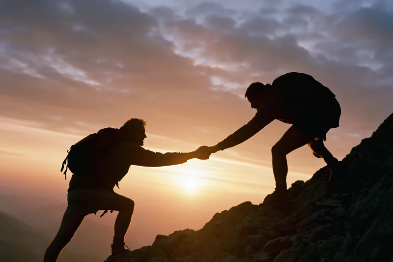 Two hikers silhouetted against a sunset, one helping the other climb a rocky mountain peak