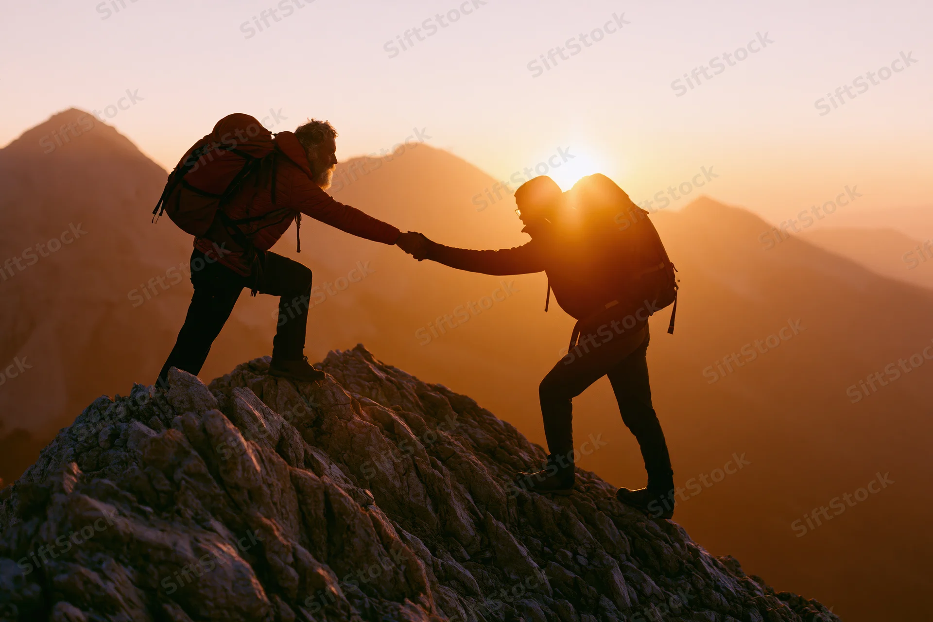 Two hikers silhouetted on rocky mountain peak at sunrise, one reaching hand to help the other climb up