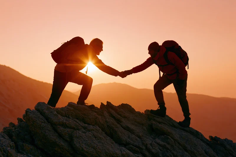 Two hikers silhouetted on rocky mountain peak reaching hands to help each other climb at sunrise