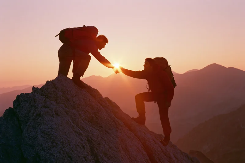 Two hikers silhouetted on rocky mountain peak reaching hands to help each other climb at sunrise