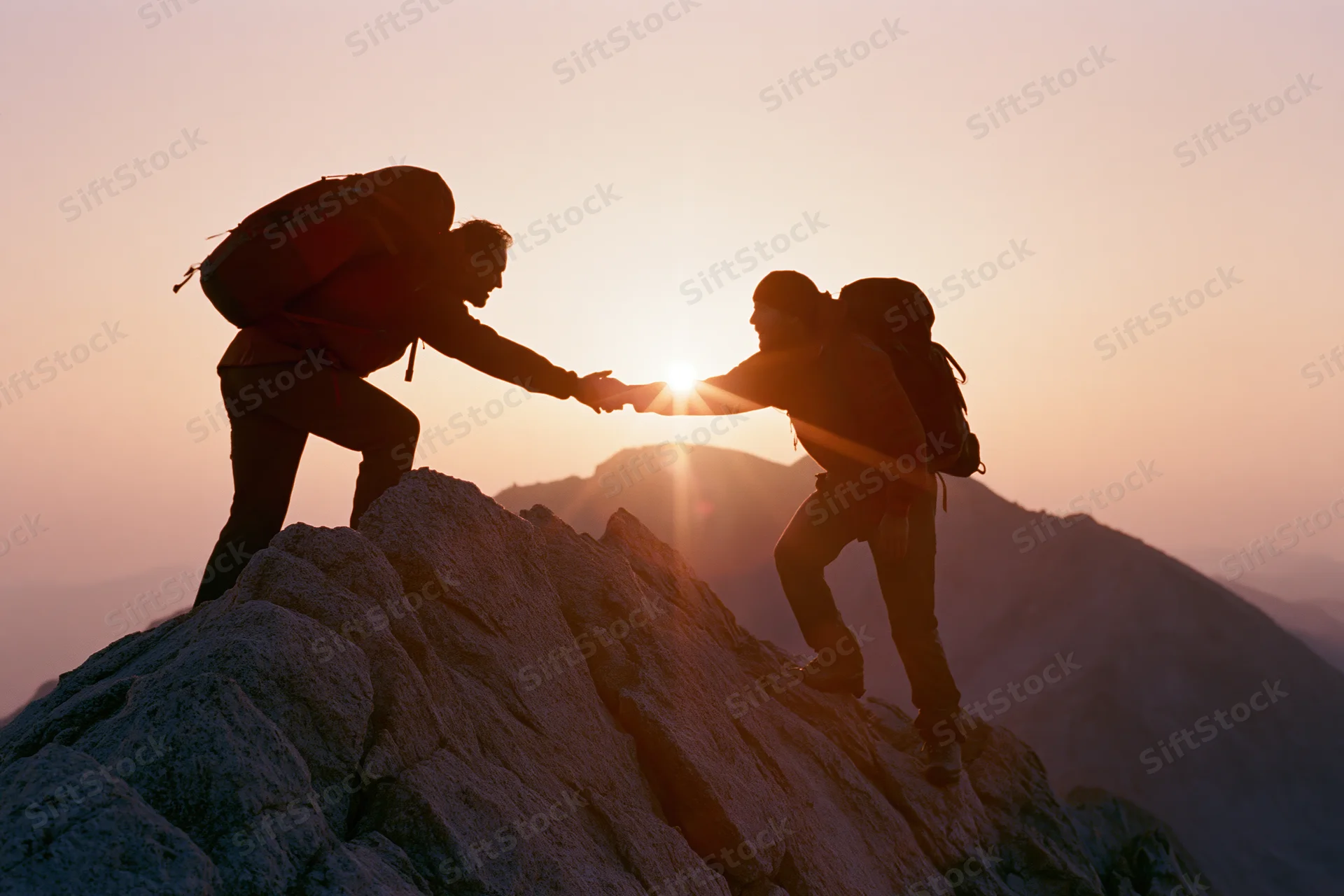 Two hikers silhouetted on rocky mountain peak reaching to help each other climb at sunset