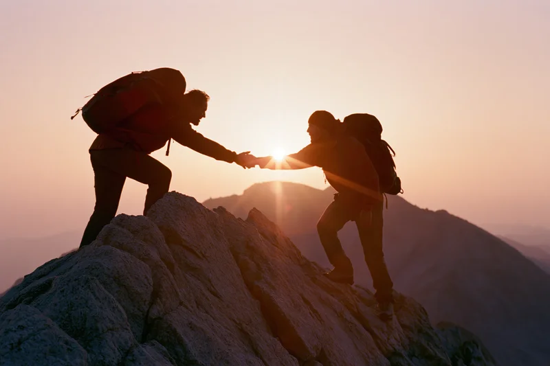 Two hikers silhouetted on rocky mountain peak reaching to help each other climb at sunset