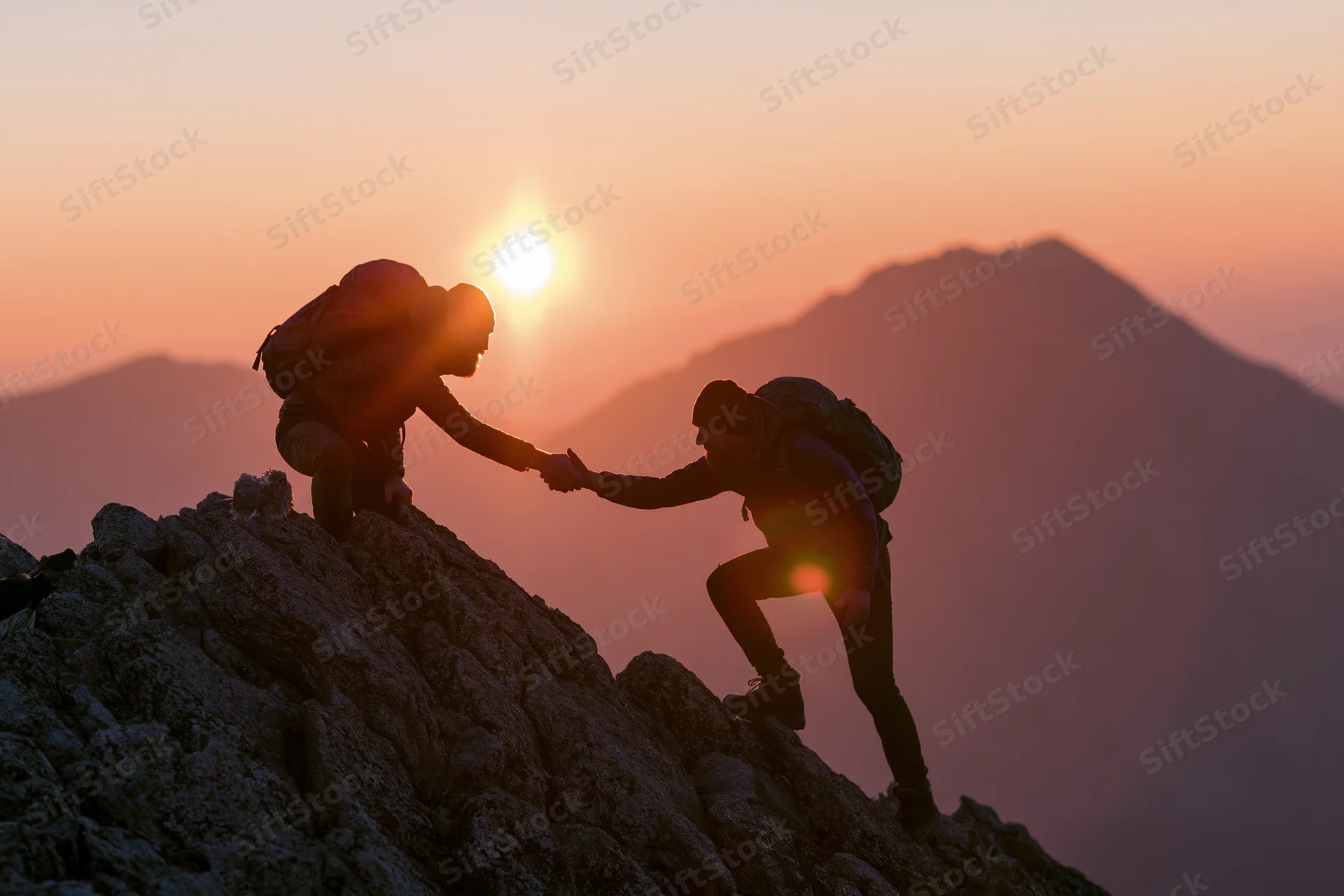 Two hikers silhouetted on rocky mountain peak at sunrise, one helping the other climb up by holding hands