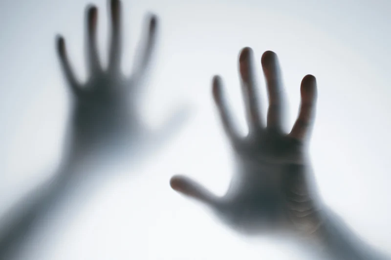 Two hands pressed against frosted glass with fingers spread and palms flat, creating blurred silhouettes against a bright background