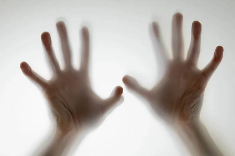 Two hands pressed against frosted glass from behind, fingers spread, palms flat, creating blurred silhouettes against a bright white surface
