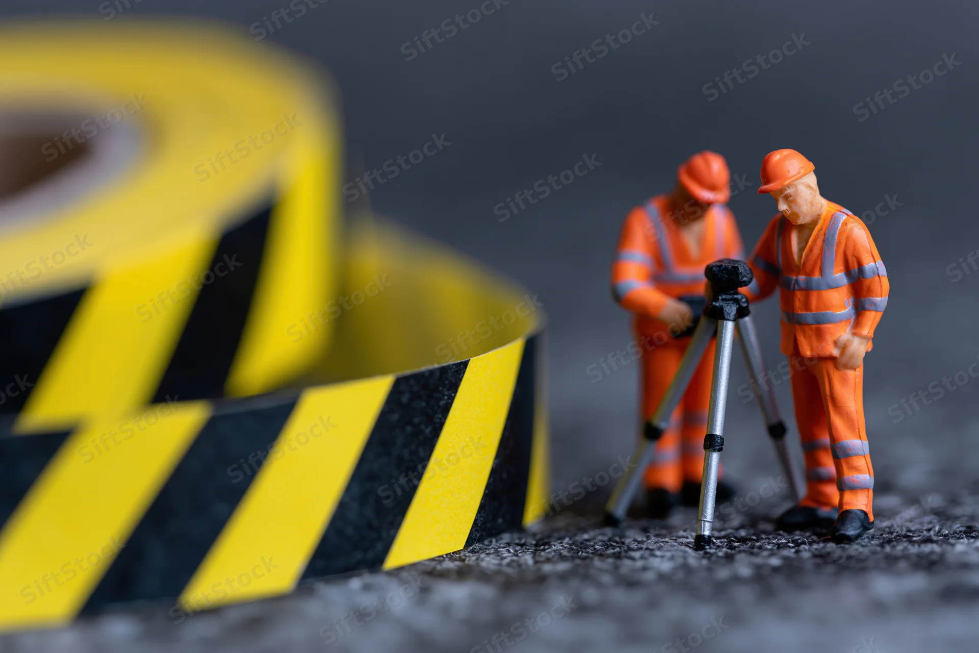 Two miniature construction workers in orange safety gear using a tripod for surveying near yellow and black caution tape on a gravel surface