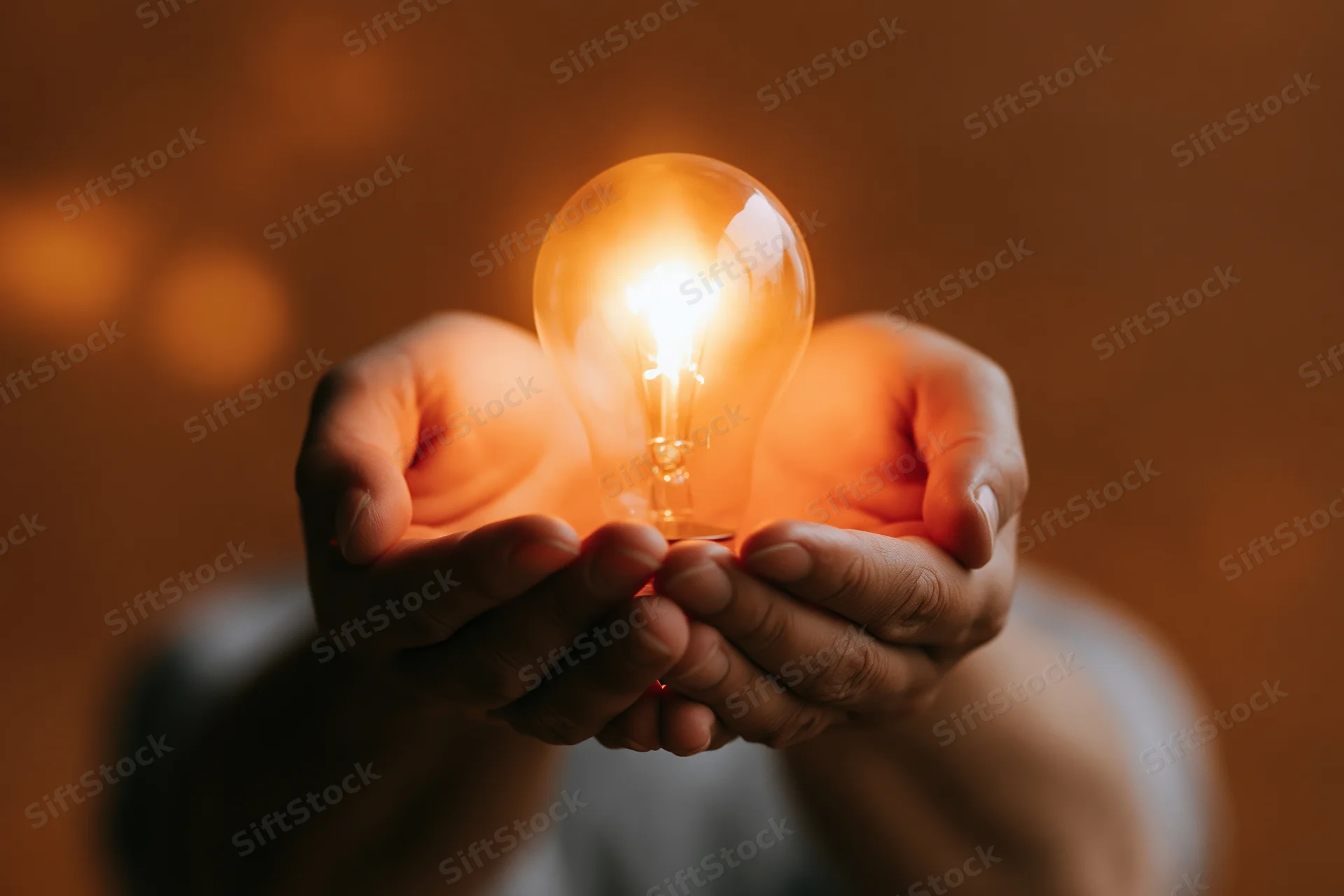 Hands cupping a glowing incandescent light bulb in closeup view