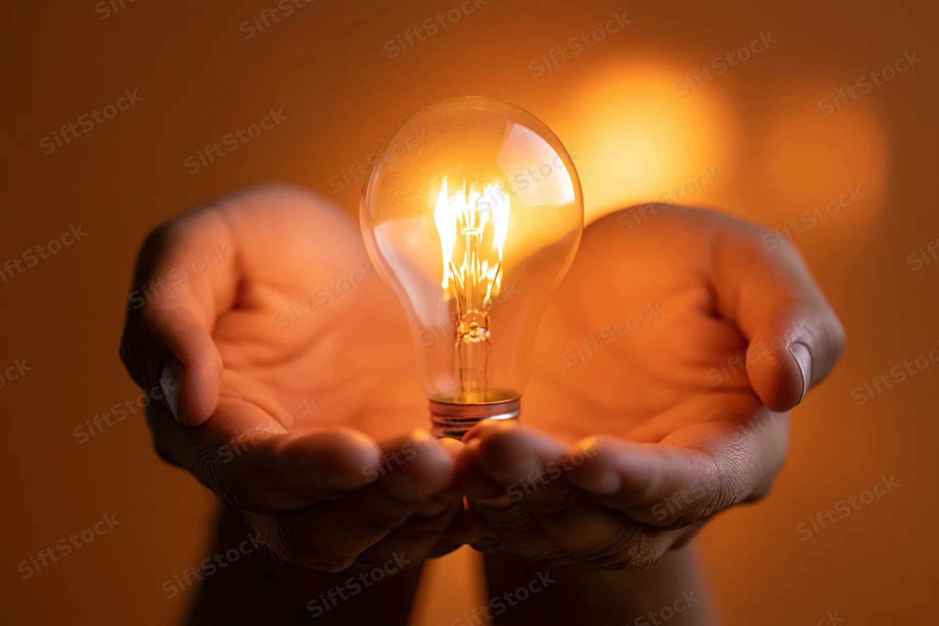 Hands cupping a glowing incandescent light bulb against a warm orange background