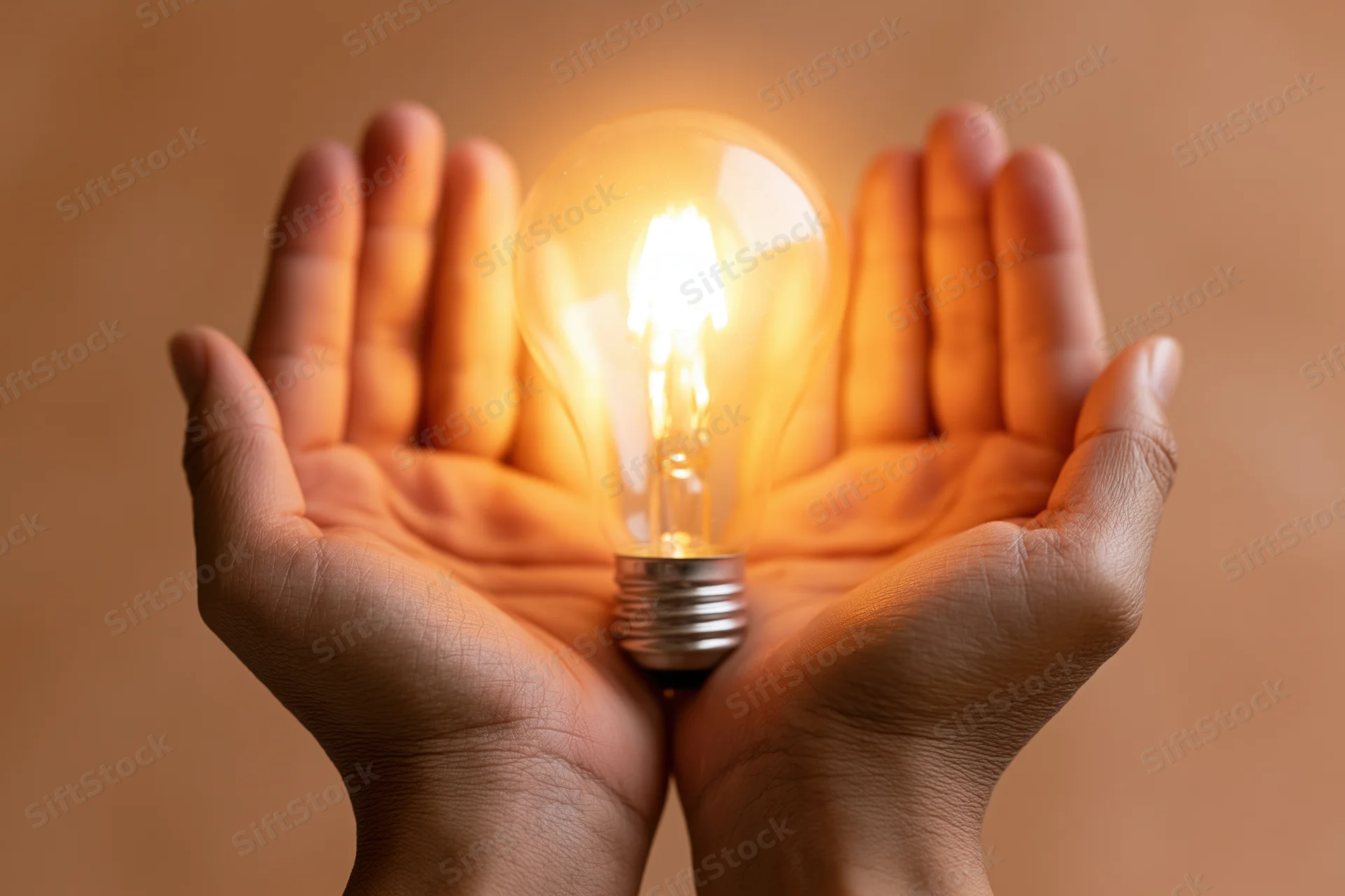 Hands cupping a glowing incandescent light bulb against a warm background