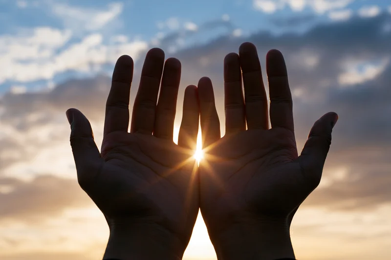 Hands raised palms-up toward the sky with sun shining through at sunrise or sunset