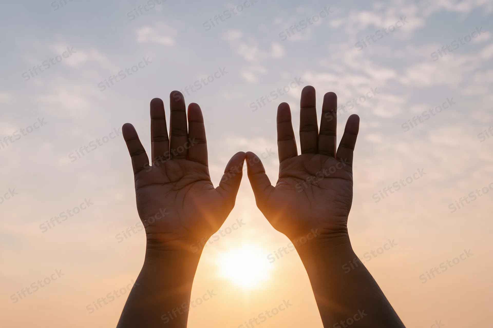 Hands raised upward framing the sun against a sunset sky