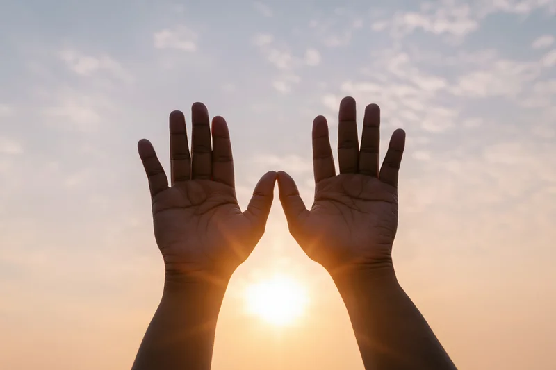 Hands raised upward framing the sun against a sunset sky