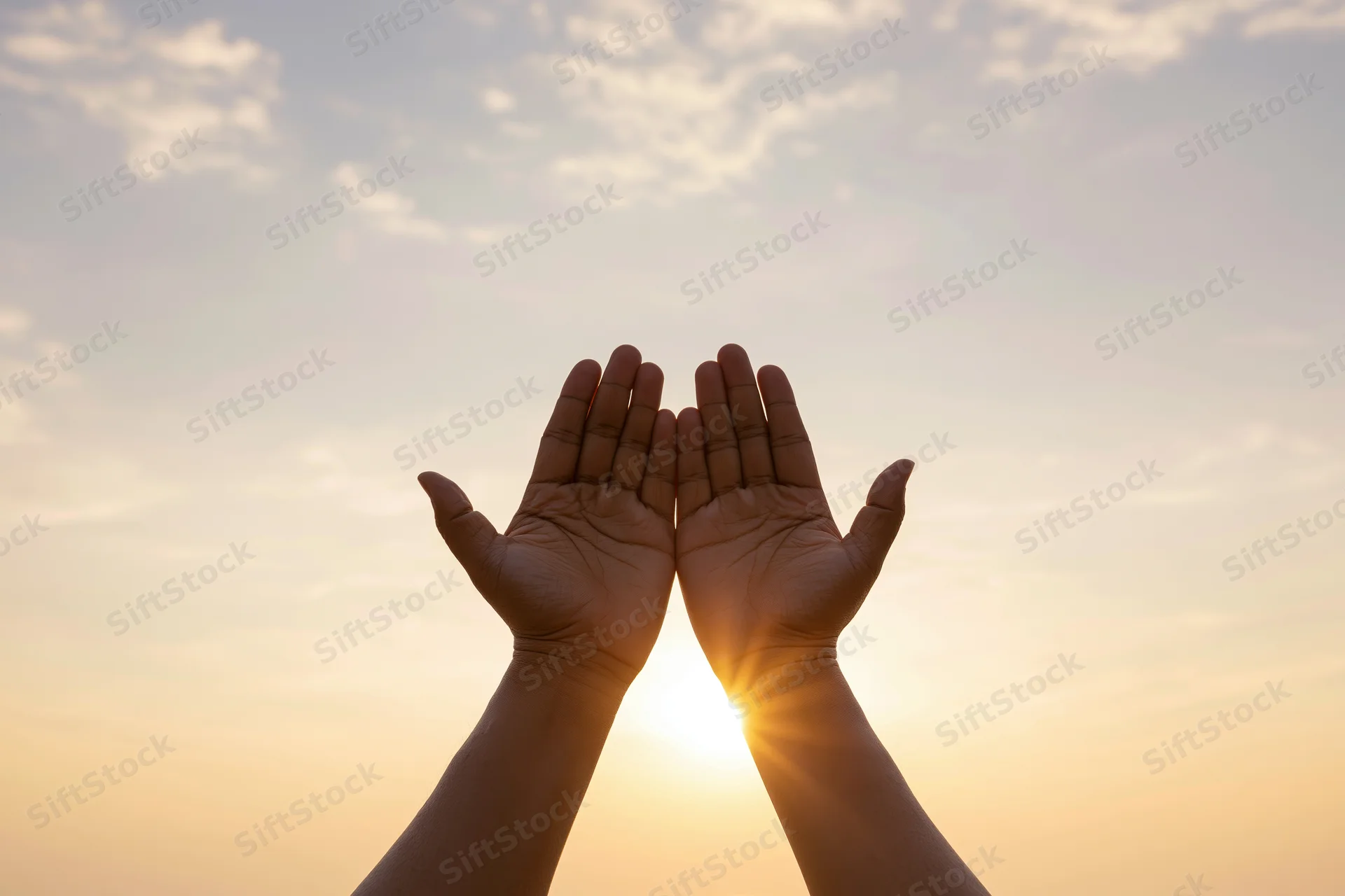 Hands raised upward framing the sun at sunset against a soft sky