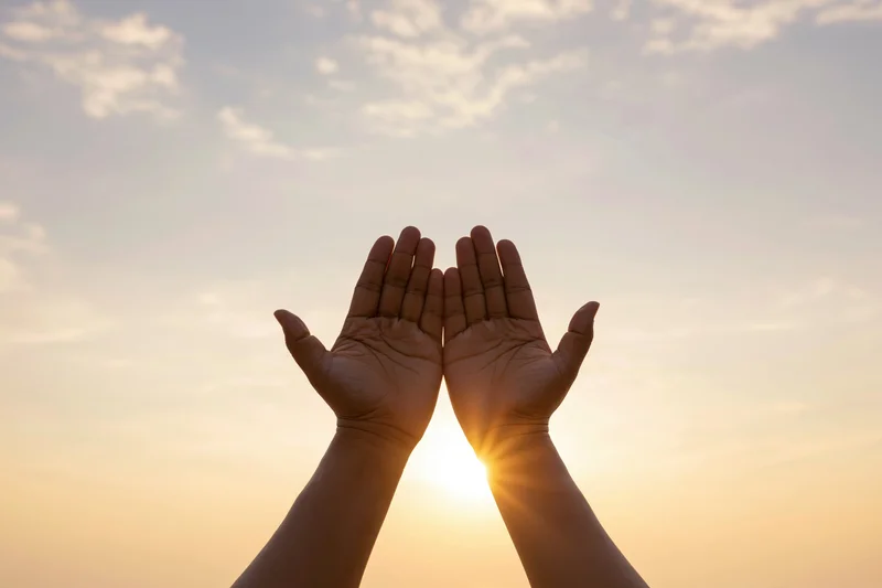 Hands raised upward framing the sun at sunset against a soft sky