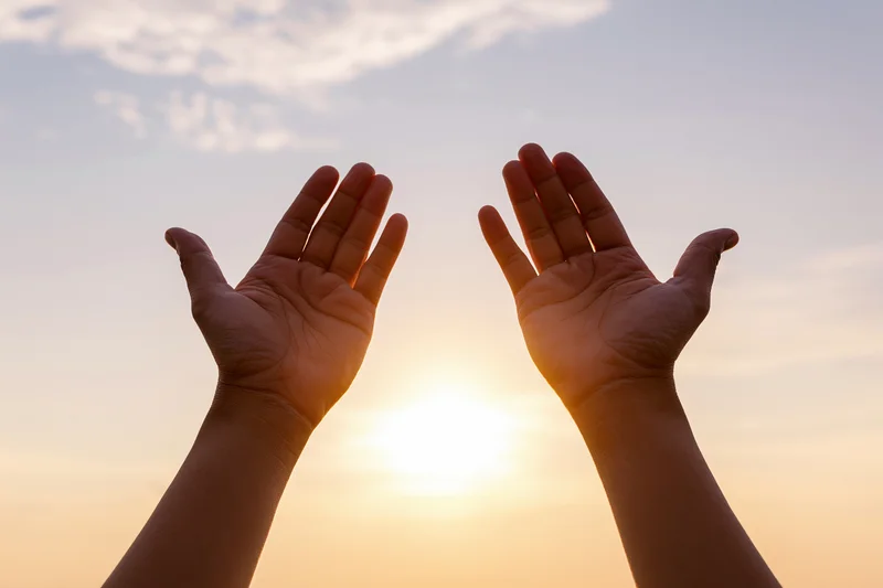 Hands raised upward against a sunset sky with sun between them