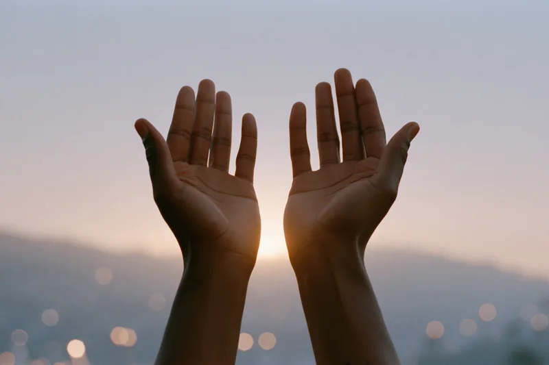 Open hands raised upward against a sunset sky with city lights in the background