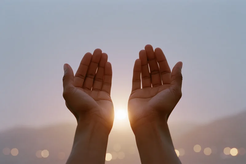 Open hands raised upward in a cupped gesture against a sunrise sky with blurred city lights below