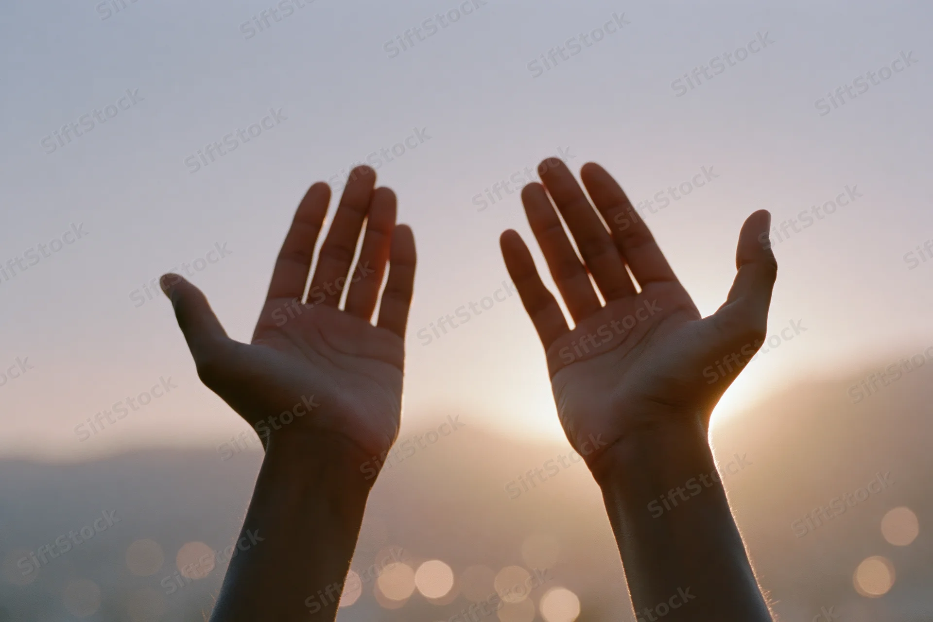 Hands raised upward in a cupped gesture against a sunset sky with blurred city lights below