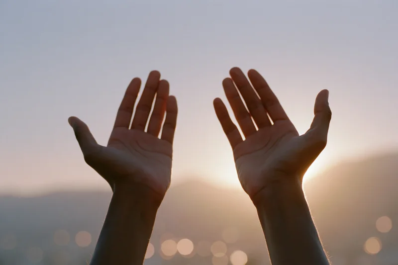 Hands raised upward in a cupped gesture against a sunset sky with blurred city lights below