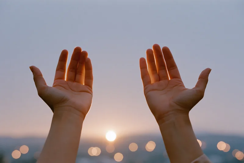 Hands raised in cupped gesture against sunset sky with city lights blurred below