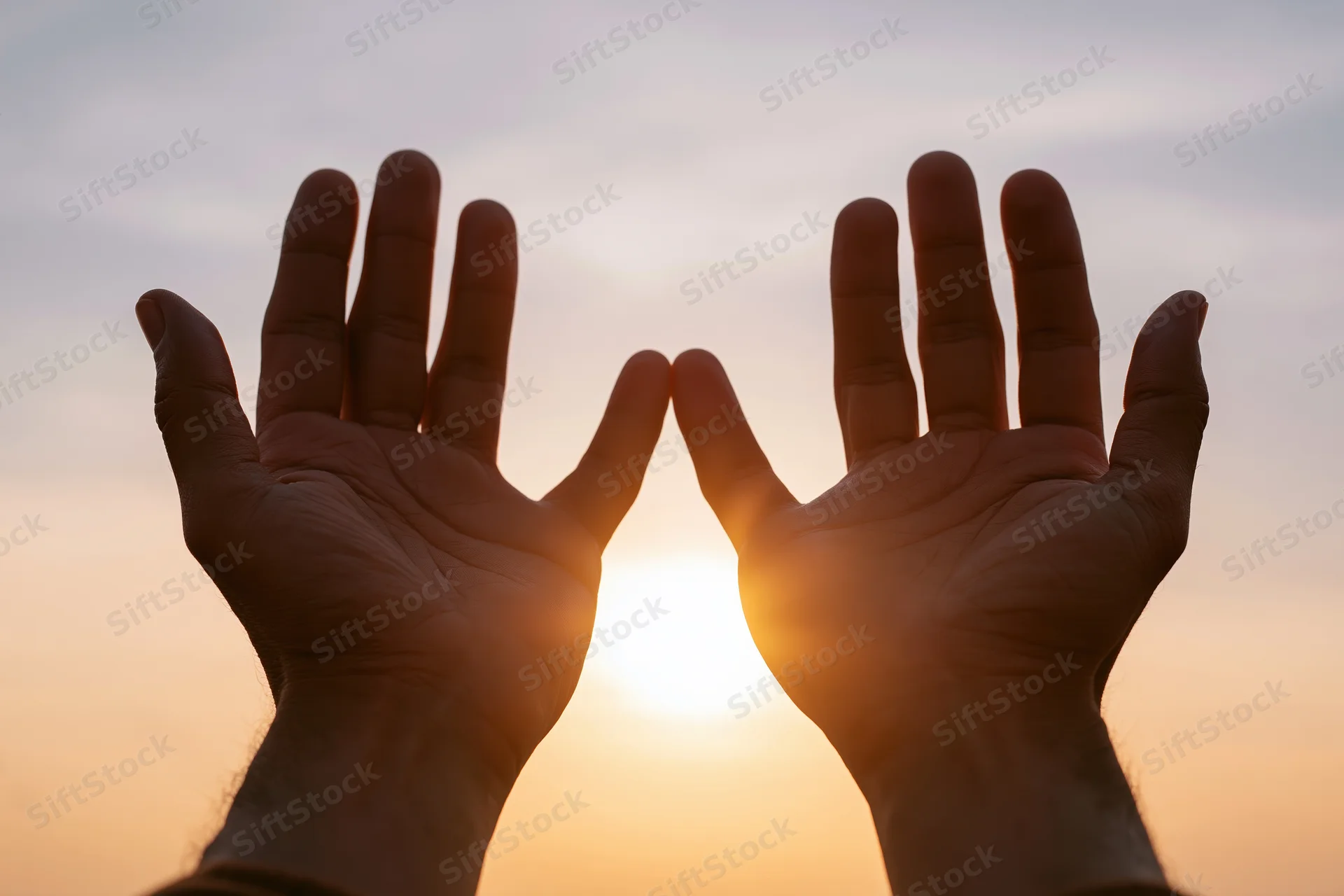 Two open hands raised toward the sky framing the setting sun at twilight