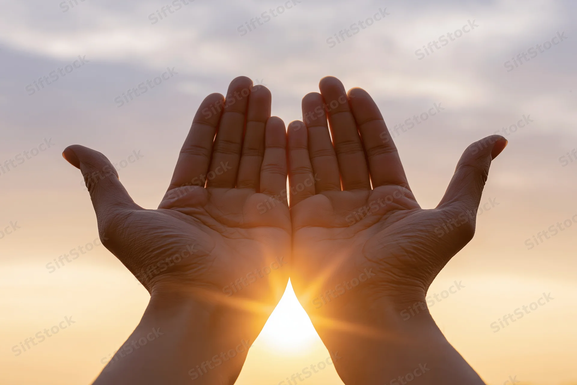 Hands raised upward framing the sun at sunrise against a soft sky