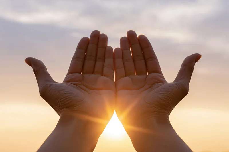 Hands raised upward framing the sun at sunrise against a soft sky