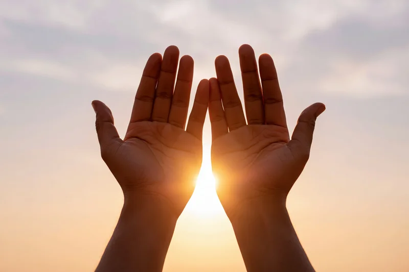 Hands raised upward framing the sun at sunset against a soft sky