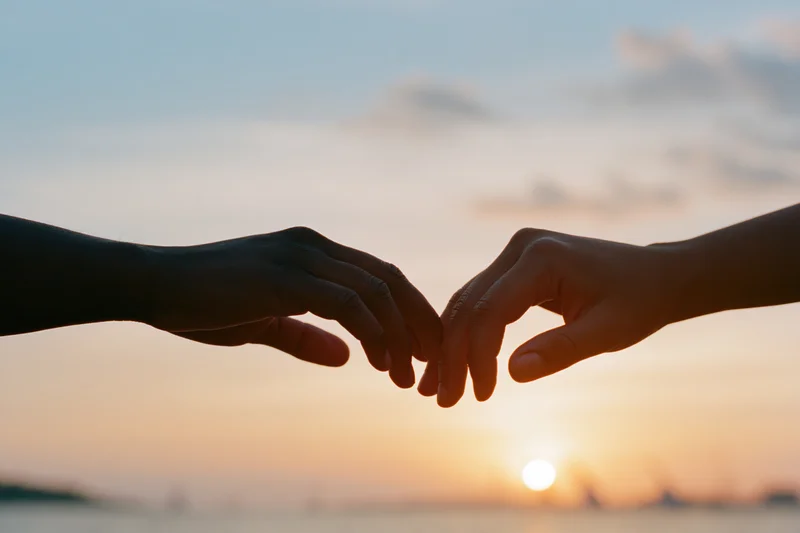 Two hands reaching toward each other in silhouette against a sunset sky