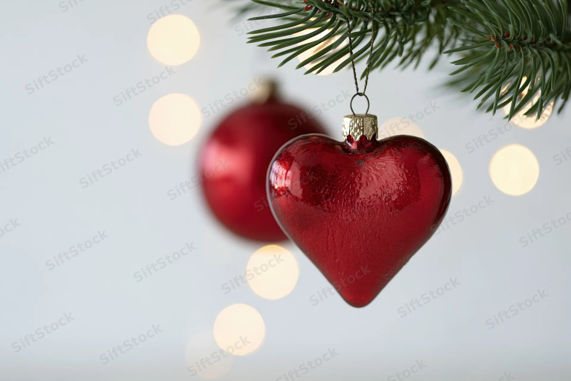 Red heart-shaped glass ornament hanging from a pine branch with blurred lights in the background