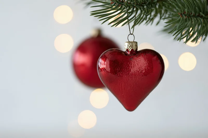 Red heart-shaped glass ornament hanging from a pine branch with blurred lights in the background