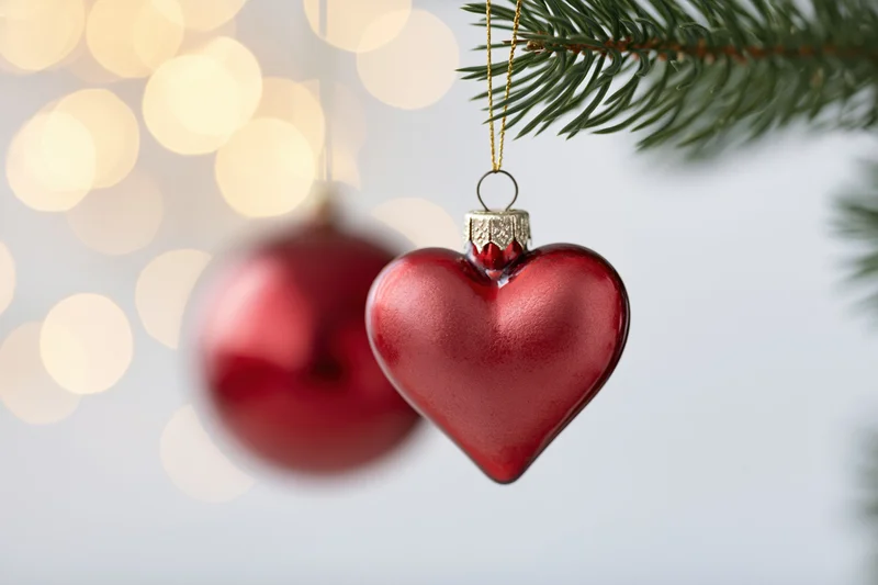 Red heart-shaped glass ornament hanging from a pine branch with blurred lights in the background