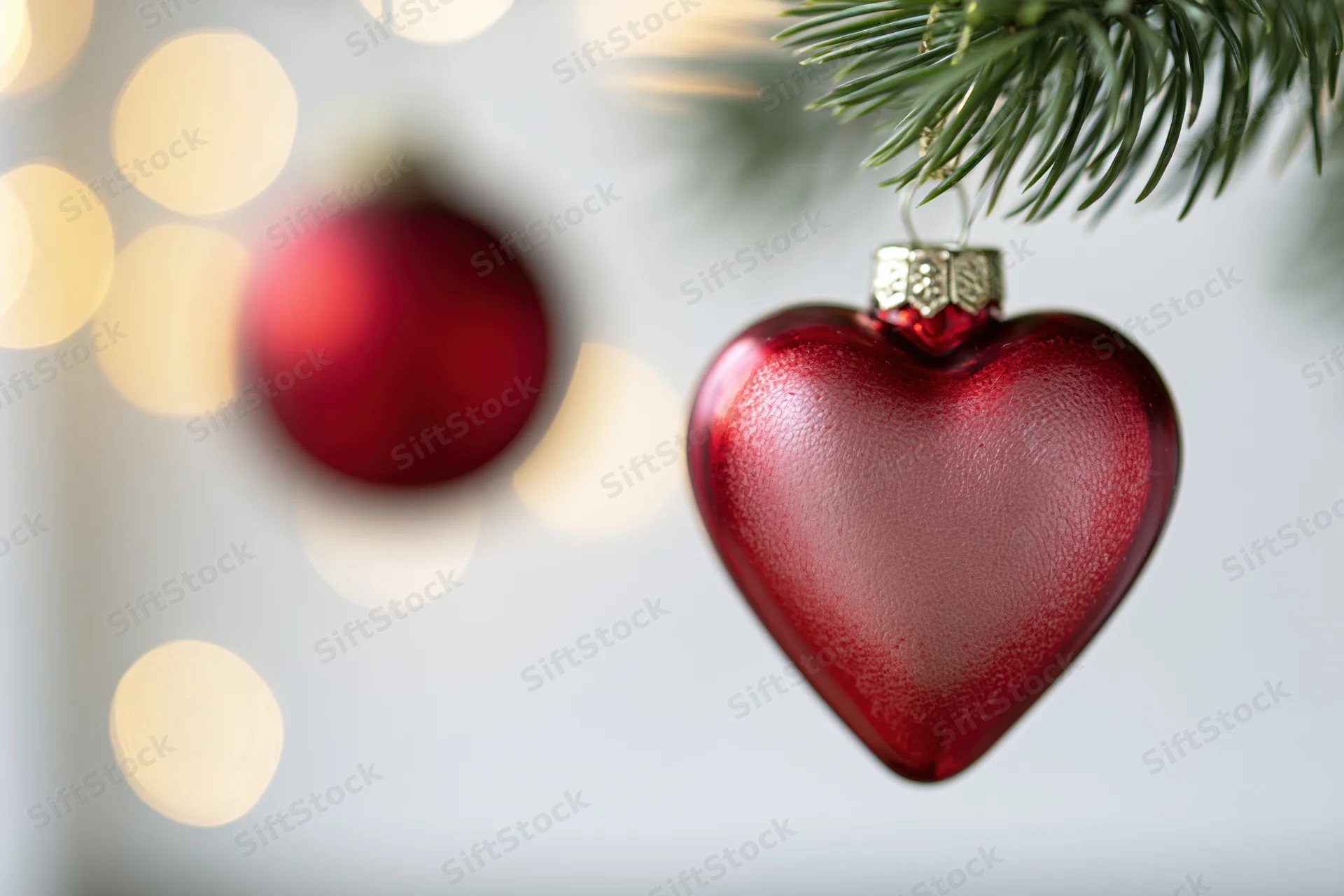 Red heart-shaped glass ornament hanging from a pine branch with blurred lights in the background