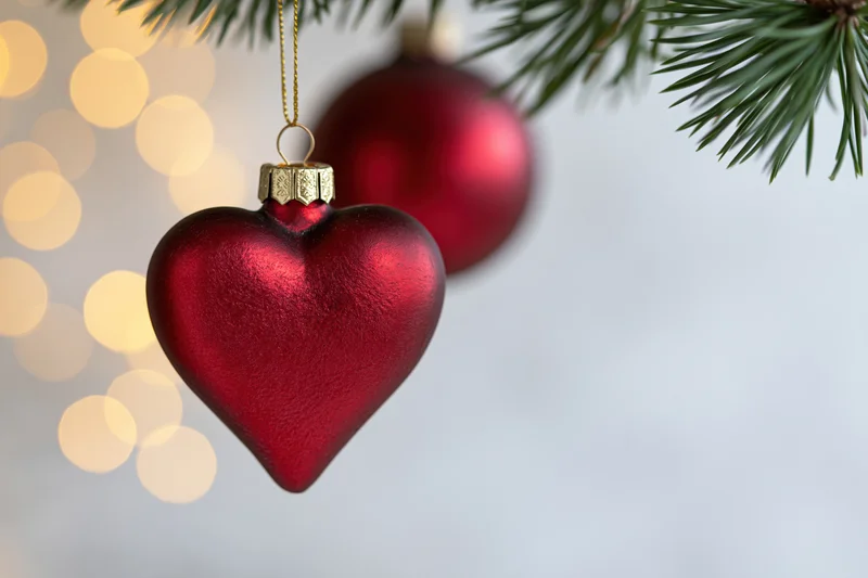 Red heart-shaped glass ornament hanging from a pine branch with blurred lights in the background