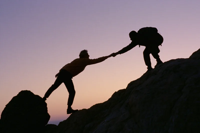 Two hikers silhouetted against twilight sky reaching hands to help each other climb rocky mountain terrain