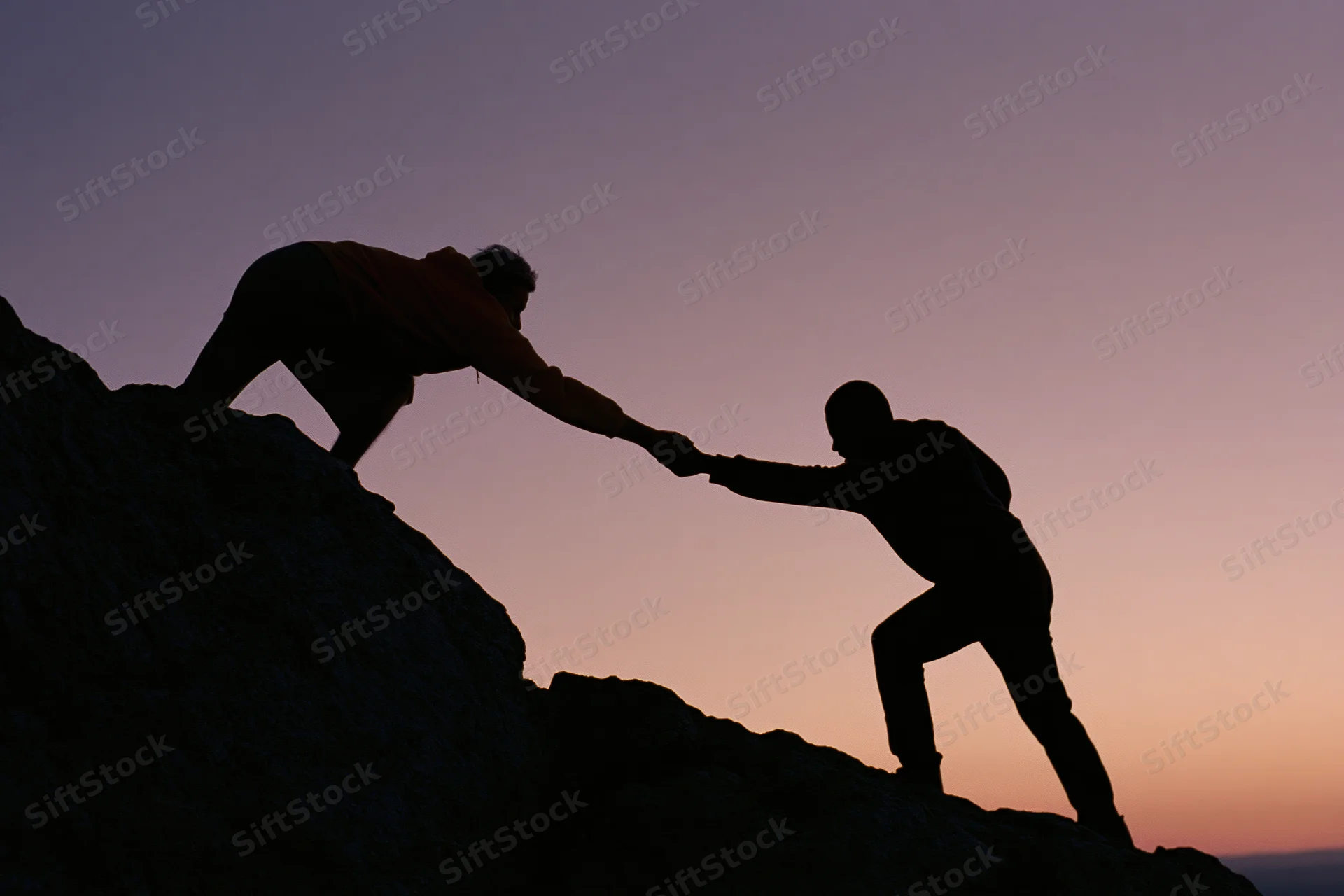 Two silhouetted climbers reaching to help each other up a rocky mountain at sunset