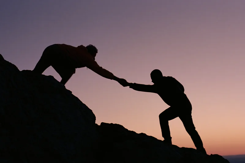 Two silhouetted climbers reaching to help each other up a rocky mountain at sunset