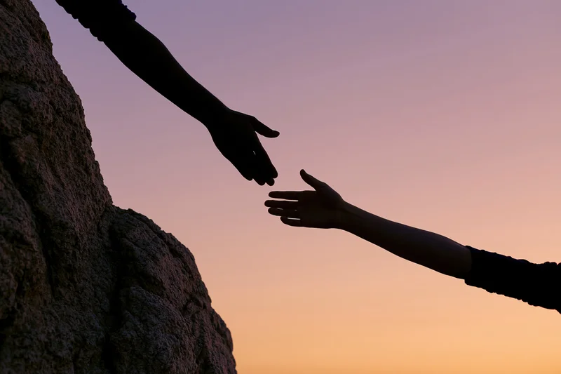 Silhouetted hands reaching toward each other against a twilight sky near a rocky cliff