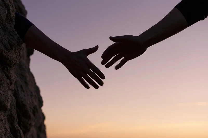 Silhouetted hands reaching toward each other against a twilight sky near a rocky cliff