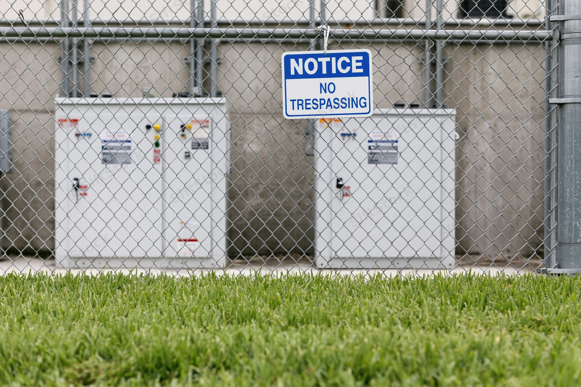 White industrial electrical utility boxes behind chainlink fence with no trespassing sign, grass in foreground