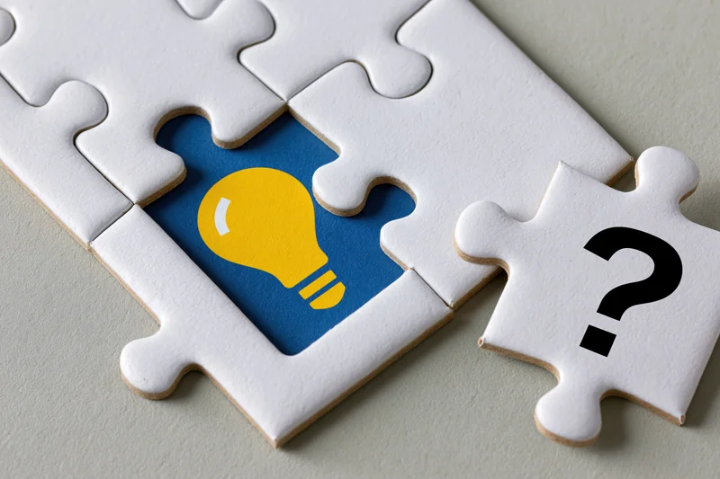 White jigsaw puzzle with missing piece revealing yellow lightbulb on blue background, one piece with question mark beside it