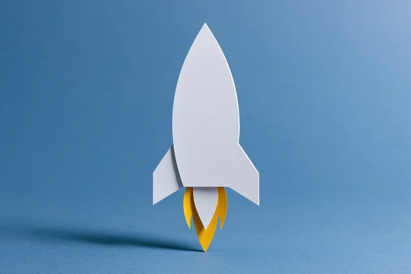 White paper-cut rocket with yellow flame cutout on blue background