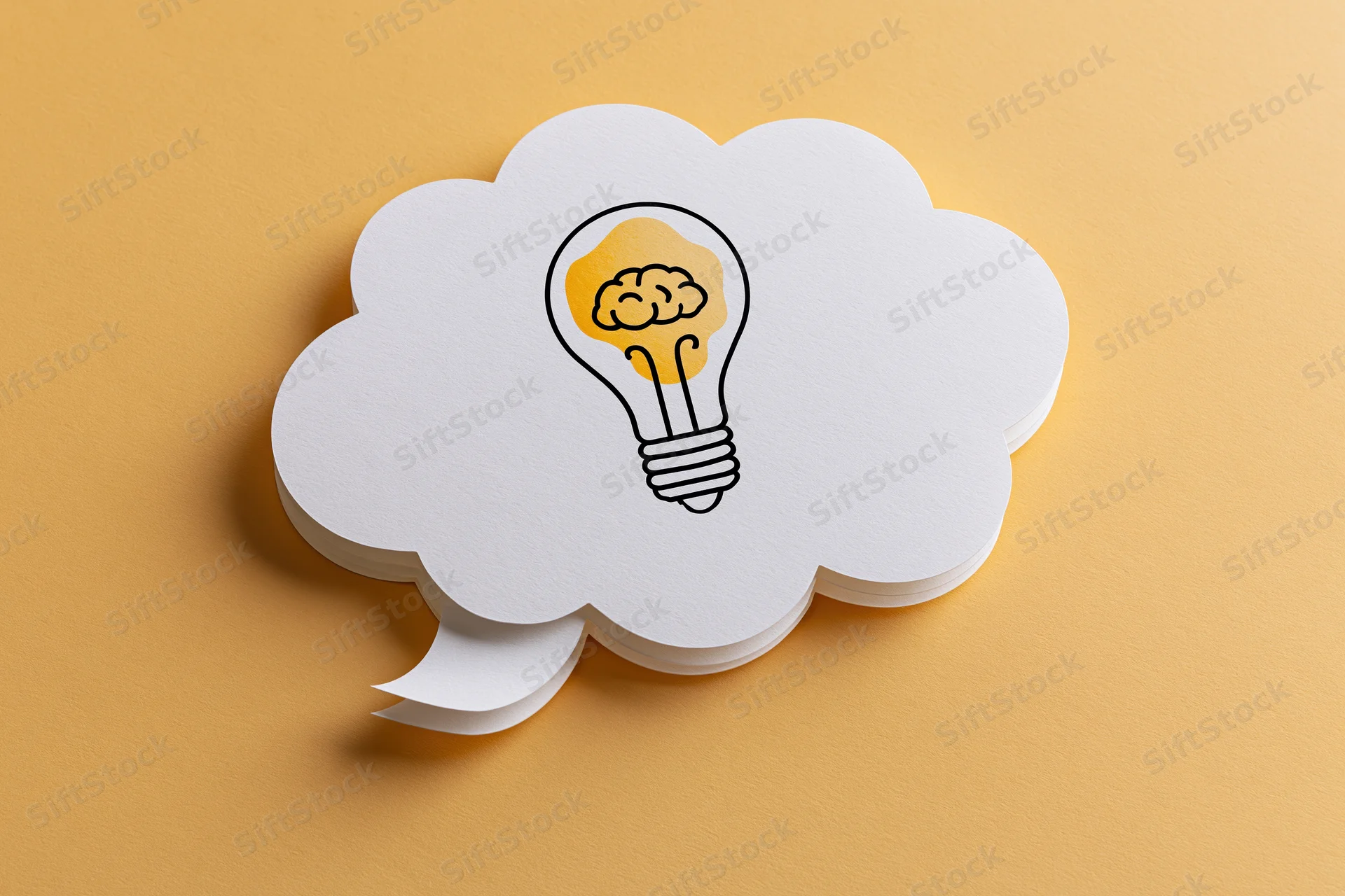 White paper thought bubble with hand-drawn lightbulb containing brain illustration on yellow surface