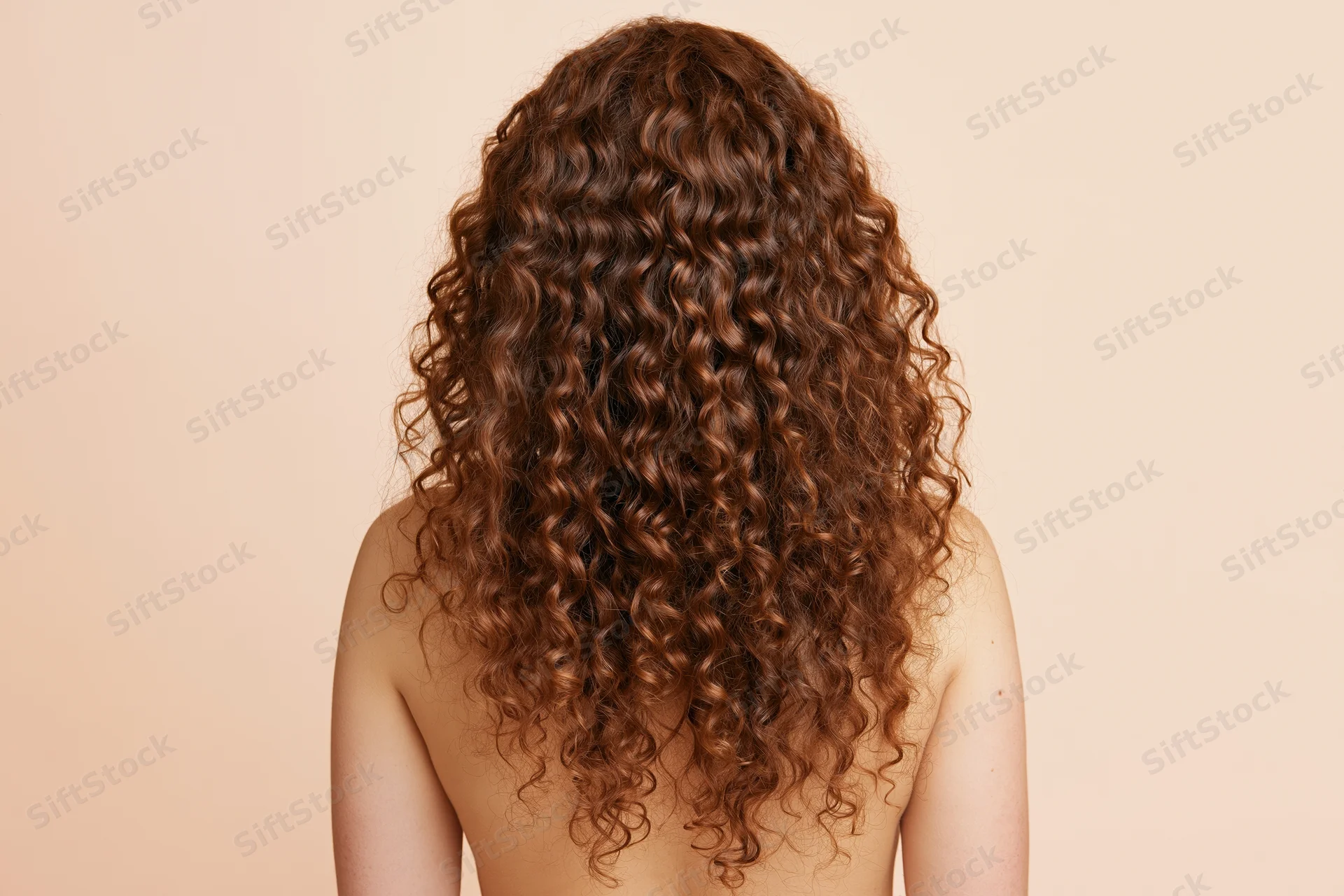 Woman seen from back with long voluminous auburn curly hair, bare shoulders, beige background