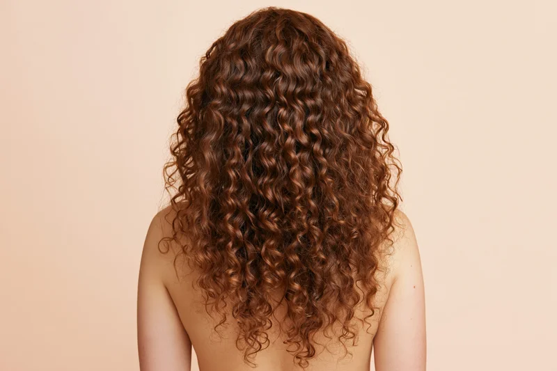 Woman seen from back with long voluminous auburn curly hair, bare shoulders, beige background