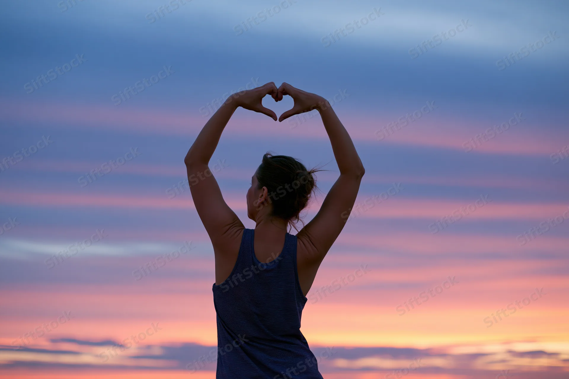 Woman from behind forming heart shape with arms against colorful sunset sky