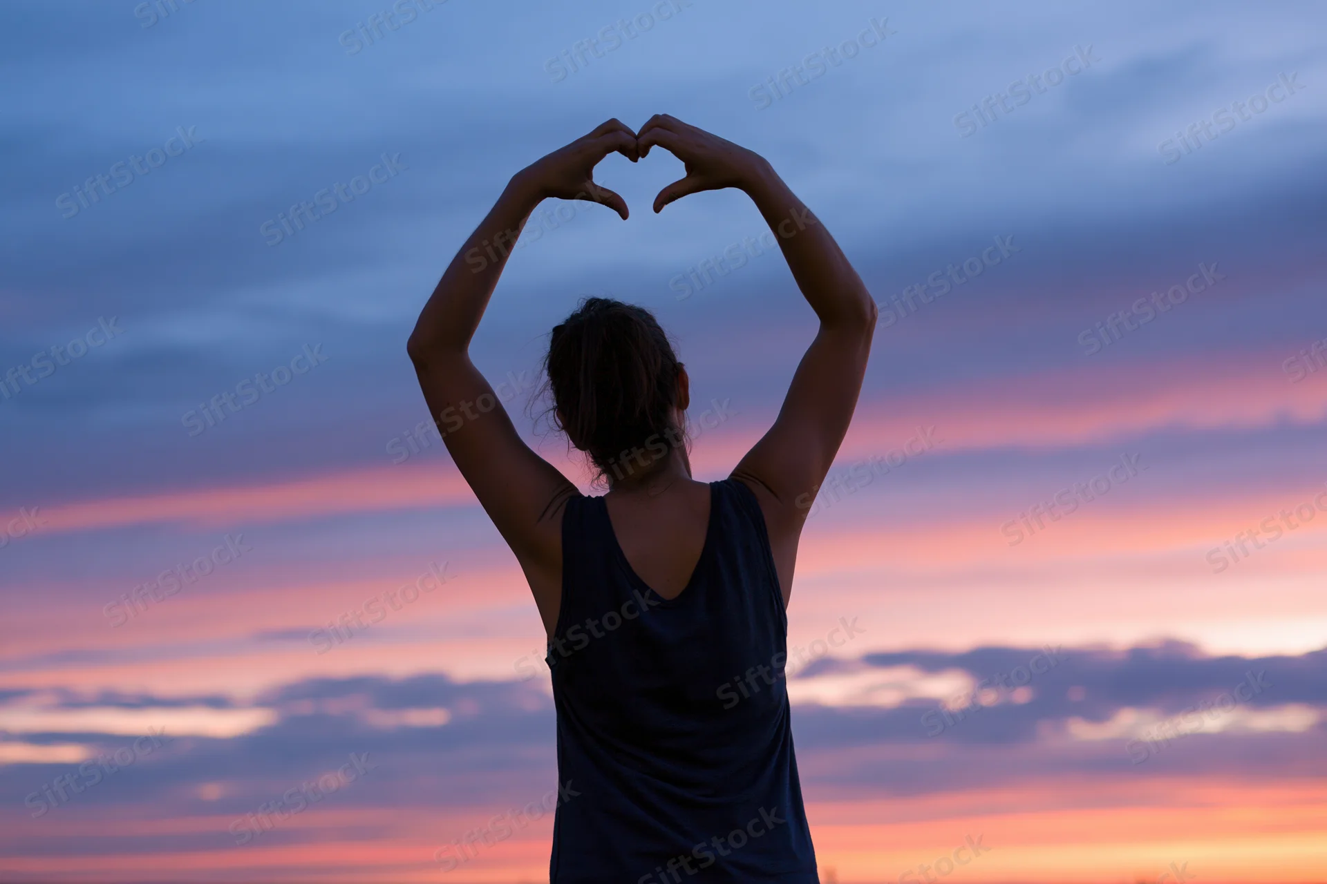 Woman from behind forming heart shape with arms against colorful sunset sky